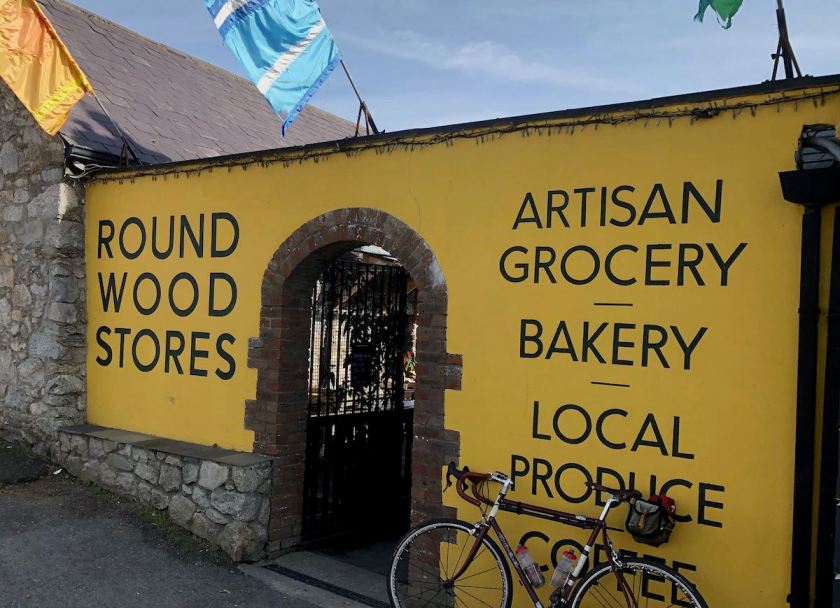 Yellow wall with arched doorway. Painted signage reads "Roundwood Stores - Artisan Grocery - Bakery - Local Produce - Coffee" in a clean, modernist sans serif font