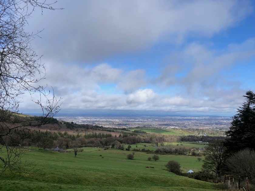 The view from Cruagh Road: green fields dropping away under partly cloudy skies, Dublin city and bay visible in the background.