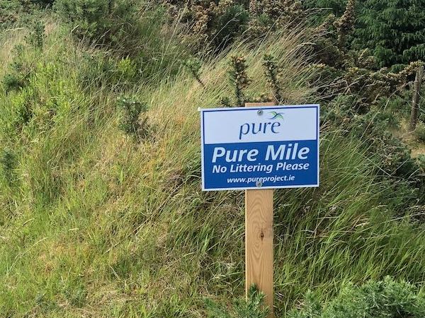 Rectangular white and blue sign reading Pure Mile No Littering Please on a post at the roadside