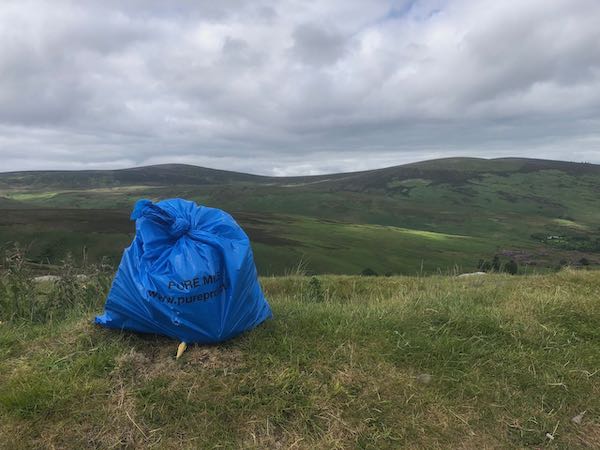 Blue plastic rubbish sack with Pure branding, standing on a ditch, mountains in the background
