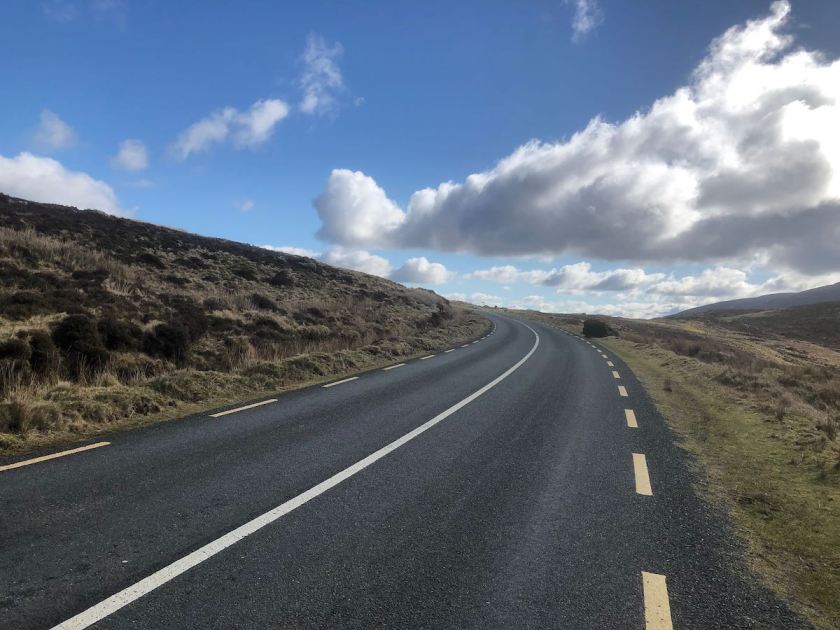 A wide road rising and swing left over a mountain pass under scattered cloud