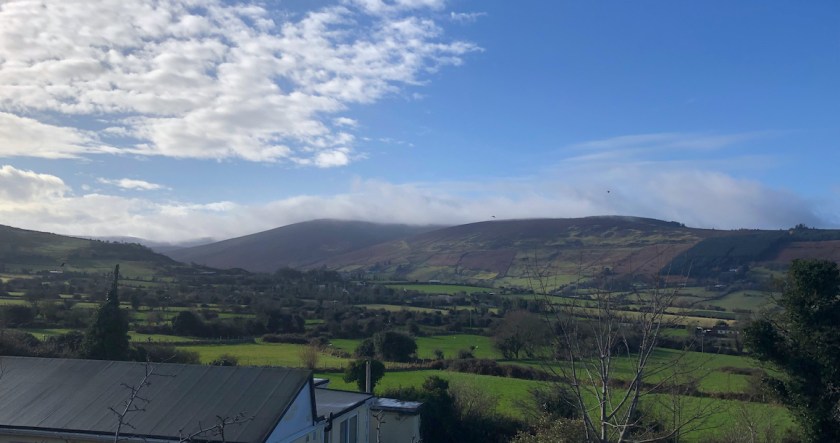 View of Glassamucky from Piperstown Road: Patchwork green fields with mountains risgin in the background.