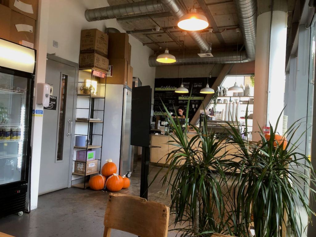 Interior of cafe, showing the counter with menu on blackboard behind it, exposed pipes in the roof, a small pile of pumpkins on the floor
