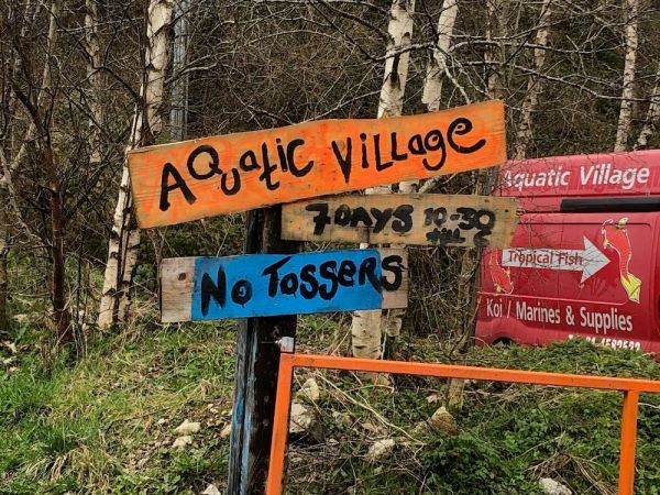 Handmade sign, painted on of planks nailed to a post: Aquatic Village, 7 days 10.30 until 6, No Tossers