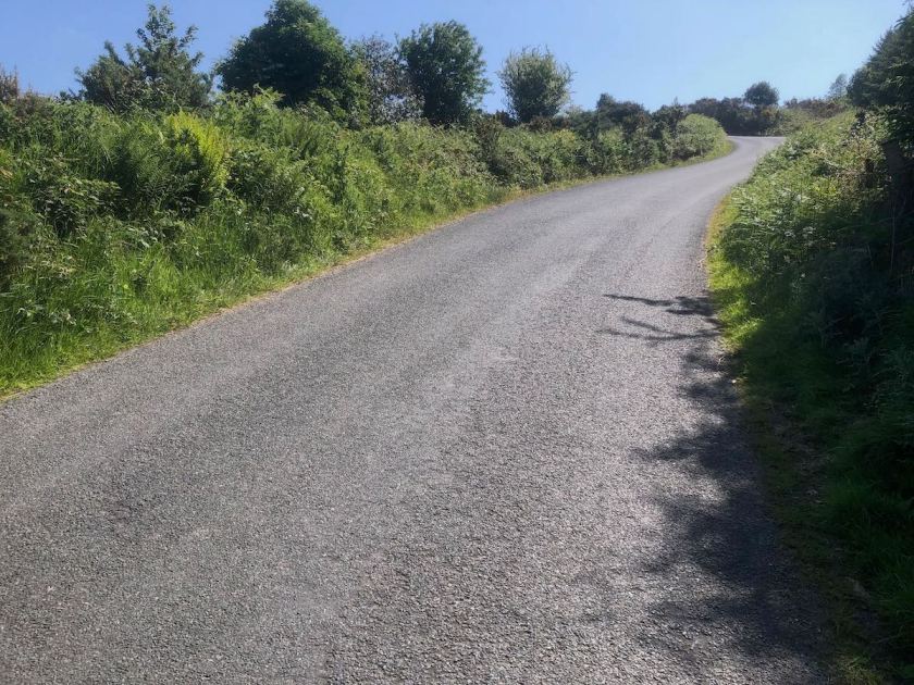 Rough country road lined with hedgerows rising steeply to a left-hand bend